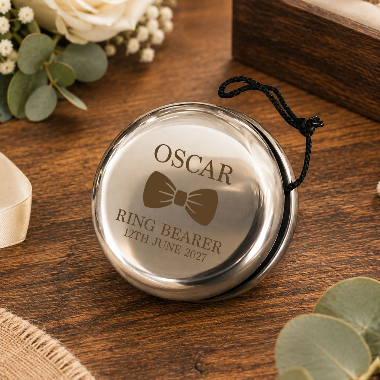 A round silver ring bearer pendant with a black hanging cord, engraved with the name OSCAR, a bow tie graphic, and the date 12th June 2027, displayed on a wooden tabletop with flowers and a ring box in the background.