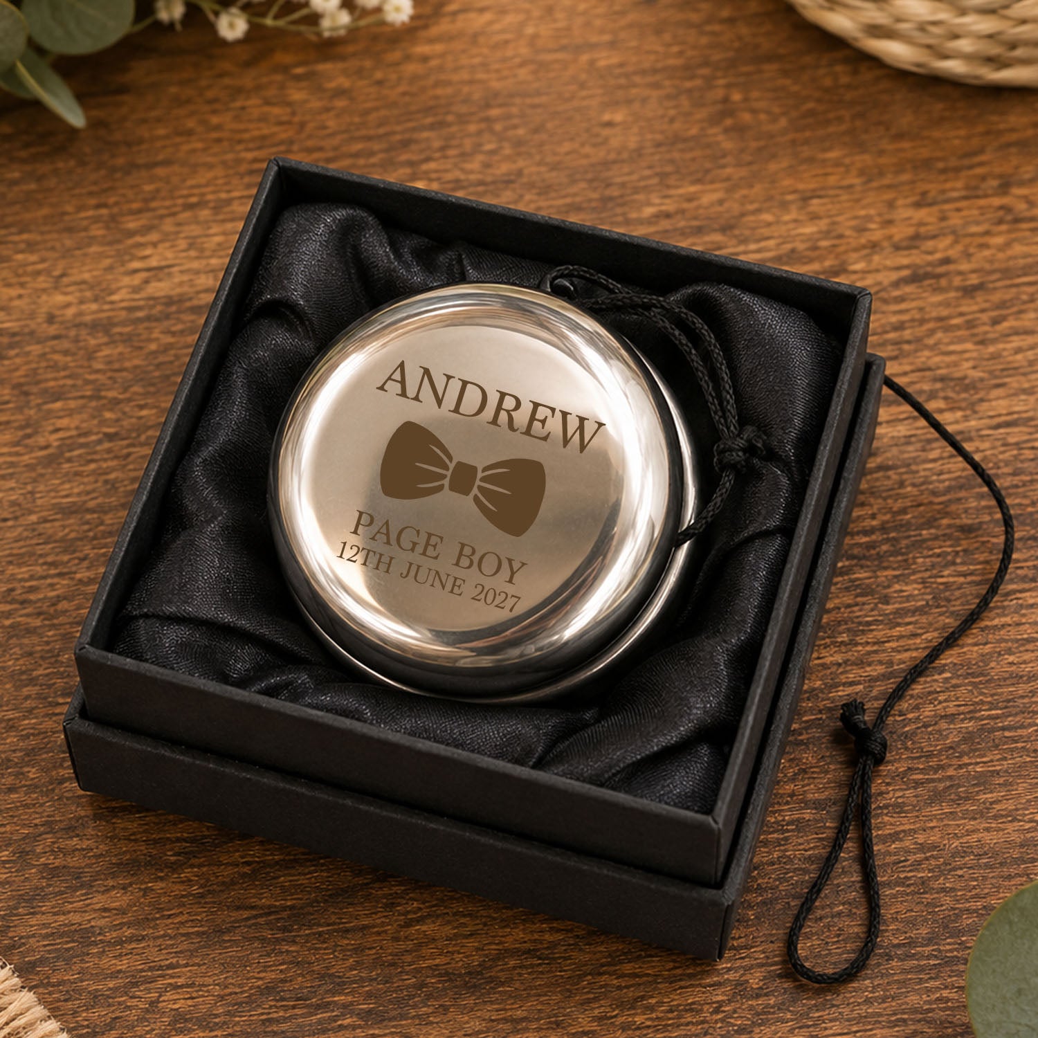 A round silver keepsake box with an engraved bow tie and the name 'ANDREW' sits in a black presentation box lined with black satin, placed on a wooden surface.