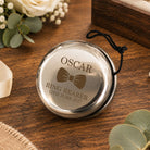 A round silver ring bearer pendant with a black hanging cord, engraved with the name OSCAR, a bow tie graphic, and the date 12th June 2027, displayed on a wooden tabletop with flowers and a ring box in the background.
