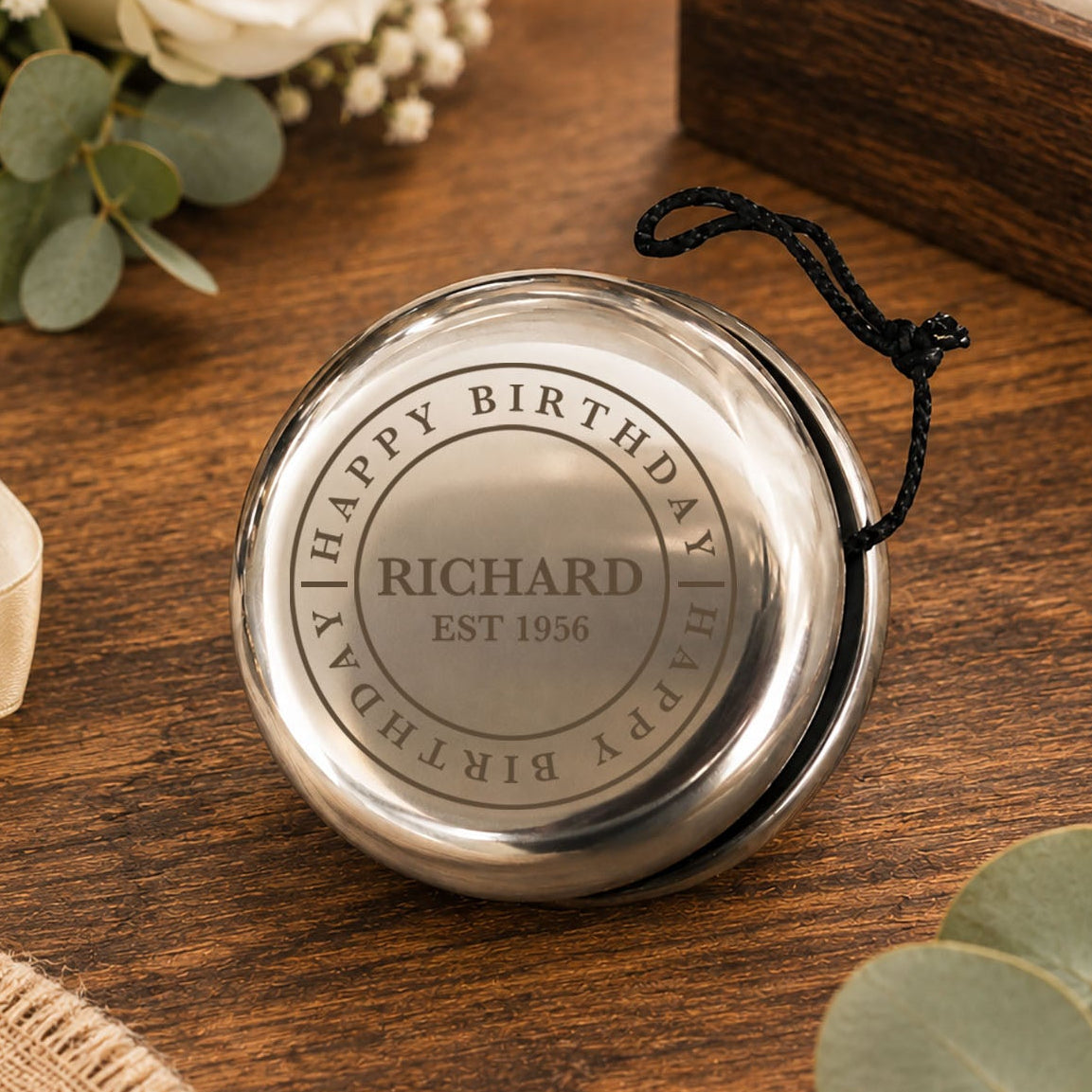 A round silver keepsake box with a black hanging cord, engraved with a heart and the message 'Jonah James Thank you for being our Page Boy xxxx', displayed on a wooden table with wedding-themed floral and ribbon props.