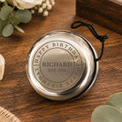 A round silver keepsake box with a black hanging cord, engraved with a heart and the message 'Jonah James Thank you for being our Page Boy xxxx', displayed on a wooden table with wedding-themed floral and ribbon props.